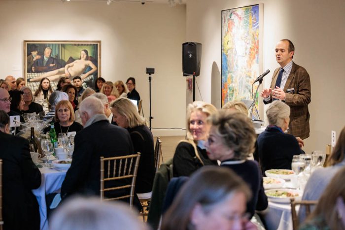 man speaking to a group of people seated at dining tables in an art gallery